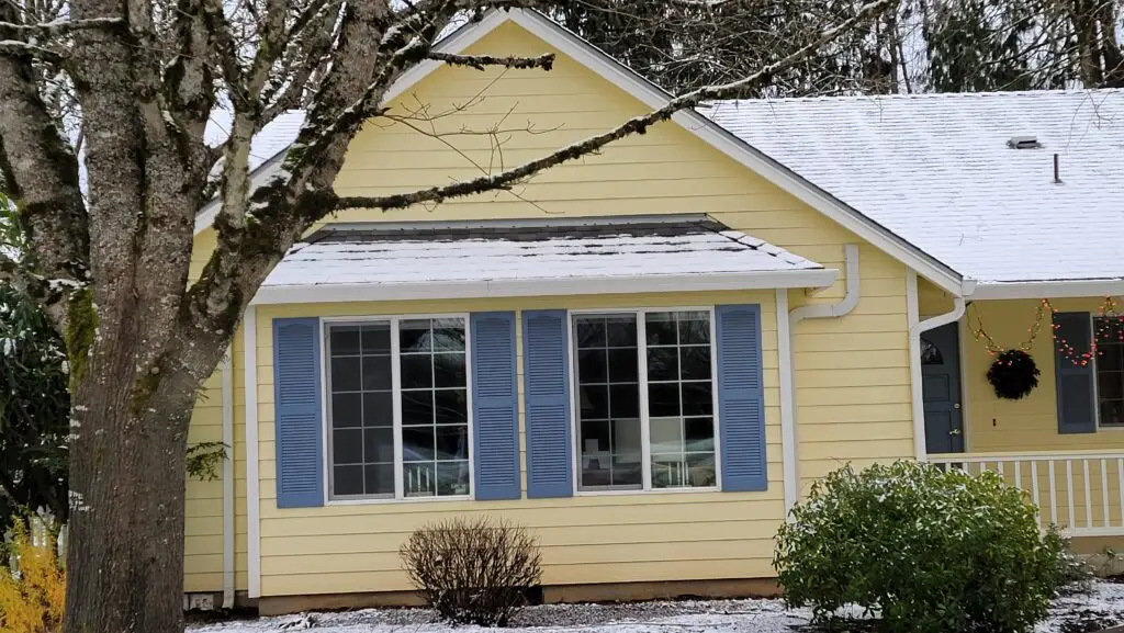 Before photo of a single story home with faded yellow siding and blue shutters prior to exterior work